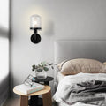 Modern black wall sconce with clear glass shade illuminating minimalist bedroom - Elirian